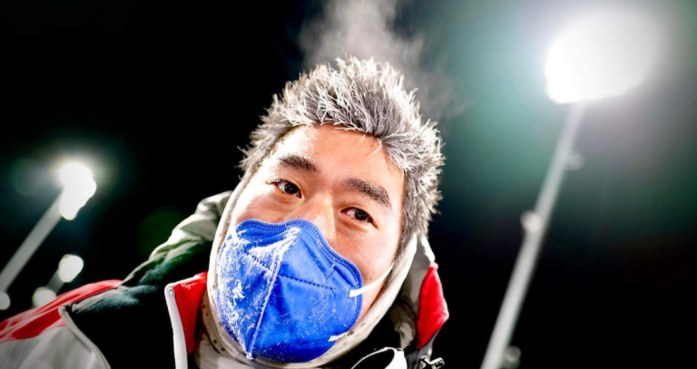 A masked man is seen with frosted hair due to the cold. Bright lights can be seen in the background. It looks like he's at an Olympic venue.